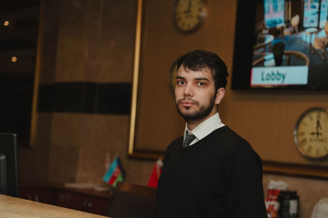 A hotel receptionist standing at the front desk in a hotel lobby, ready to assist guests.
